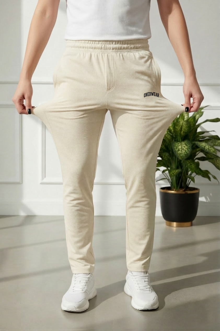 Person wearing beige pants with a logo, standing indoors next to a plant.